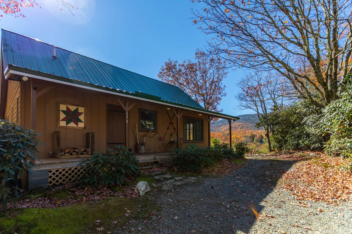 A cozy, inviting entryway featuring a wooden front door, decorative star, stack of firewood, lush green foliage, and a mountain view in the distance. The firewood hints at evenings by the fire, while the vista speaks to the broader natural surroundings.