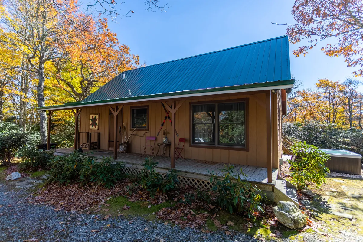 The image shows the charming exterior of a farmhouse-chic single family house, featuring a welcoming wooden porch and a distinctive green metal roof, nestled among vibrant autumn foliage. The front door boasts a unique star design, and the porch invites relaxation with ample space for gathering and enjoying the mountain scenery.