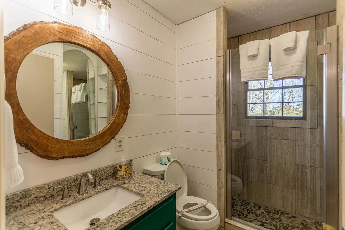 A stylish bathroom featuring a round wooden mirror above a granite countertop and a glass-door, tiled shower. The calming decor and clean finishes evoke a spa-like environment.