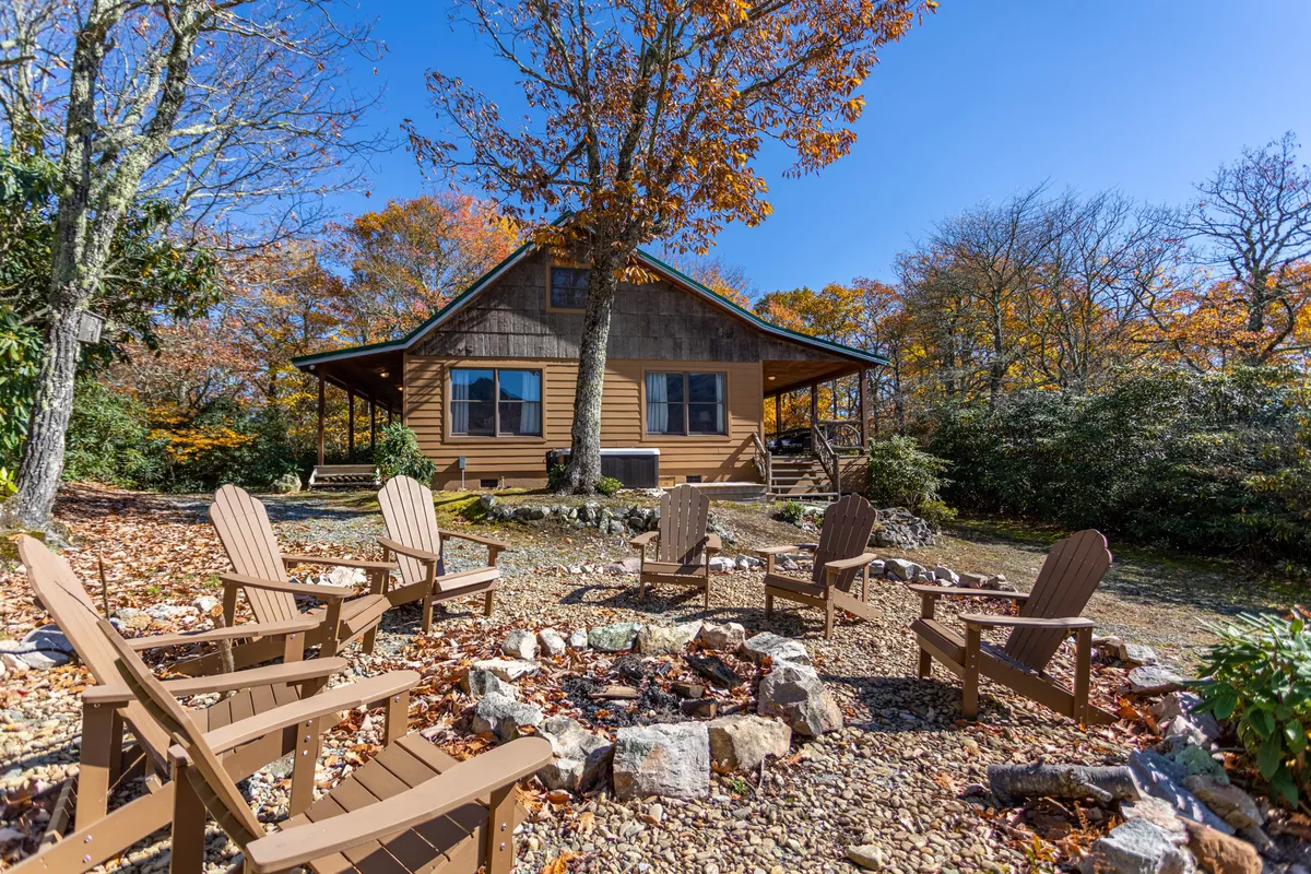 A welcoming exterior view of the cabin with a cozy fire pit, wooden chairs, and lush trees. The scene invites guests to relax after a day of adventure, enjoying campfire gatherings and outdoor relaxation.