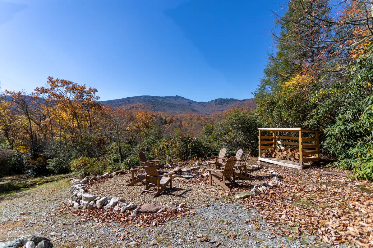 A cozy fire pit area surrounded by Adirondack chairs overlooks a stunning mountain vista, with vibrant autumn foliage in the background. The scene invites campfire gatherings, s'mores, and quiet evenings under the stars, while also showing ample open yard space for pets.
