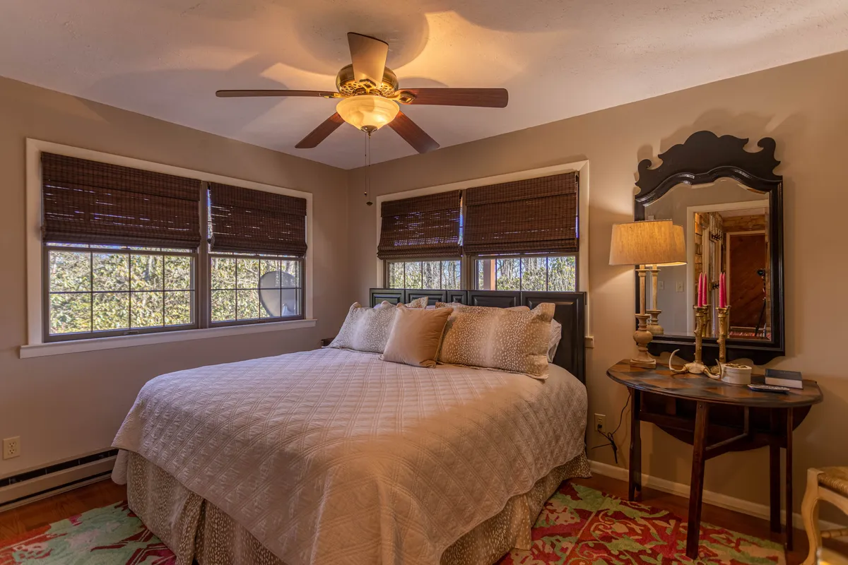 This image presents a warmly decorated bedroom featuring a king-size bed, a ceiling fan, a bedside table with a lamp, and a mirror. Natural light streams in, creating a cozy and inviting atmosphere ideal for relaxation.