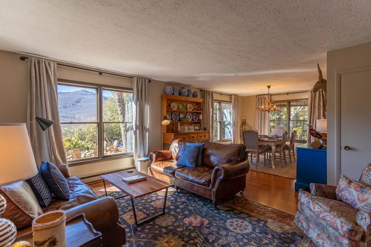 A cozy open-concept space featuring plush leather sofas arranged around a central wooden dining table, with colorful wall plates adding a touch of farmhouse style. Large windows allow plenty of natural light and offer stunning mountain views in the background.